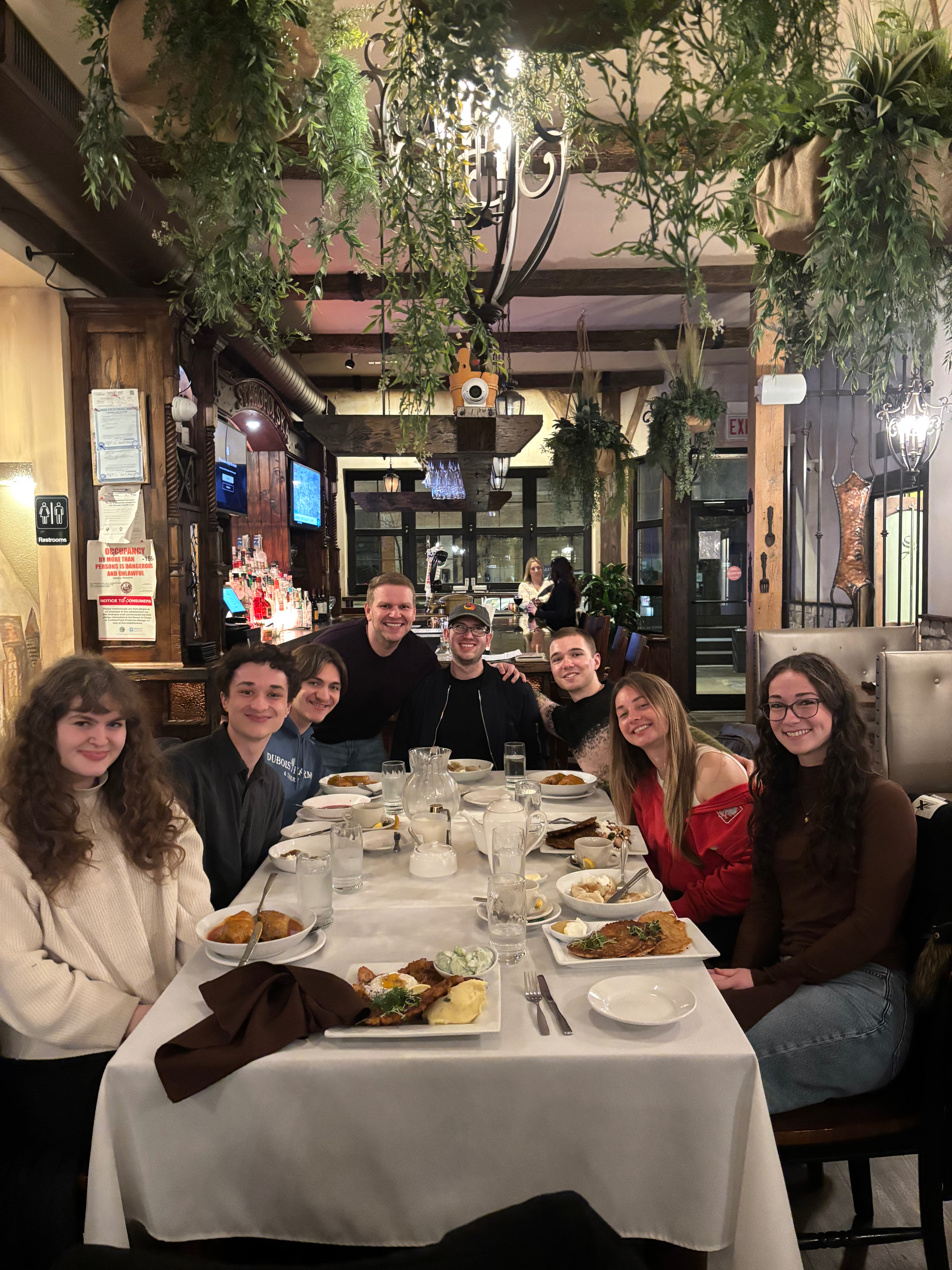 Seven undergraduate students and 1 professor sit around a restaurant table with many green plants hanging suspended from the ceiling